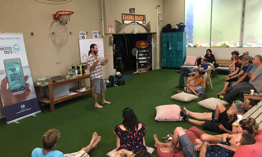 A researcher giving a jellyfish talk to participants seated on cushions at a beach club, beside a Meduzot Ba'am project banner