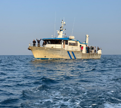 A white research vessel at sea with people waving from the deck
