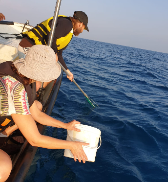 Two researchers sampling from a small boat on open blue water, one holding a bucket and one a sampling pole