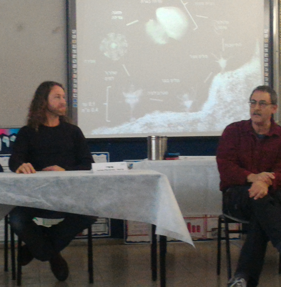 Two scientists seated before the class in discussion, with a Hebrew diagram of the jellyfish life cycle projected behind them