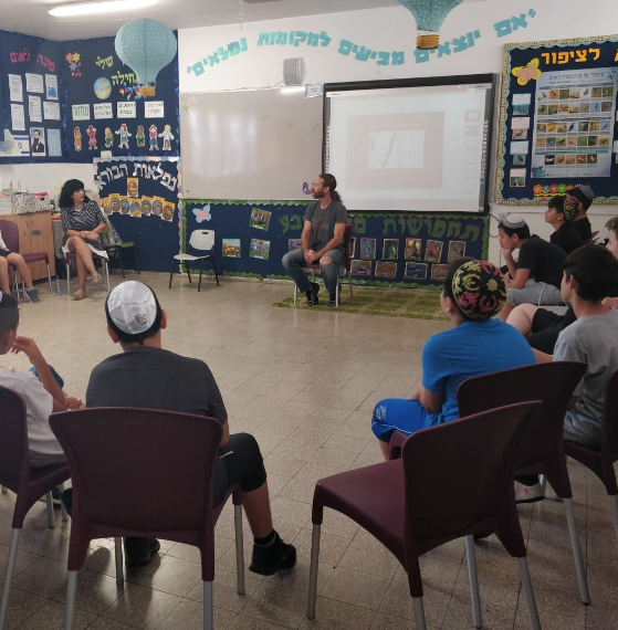 A researcher sitting in a circle with elementary school pupils in a classroom, a presentation projected behind him