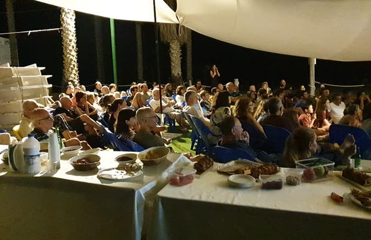 A large seated audience under a canopy at an evening community lecture